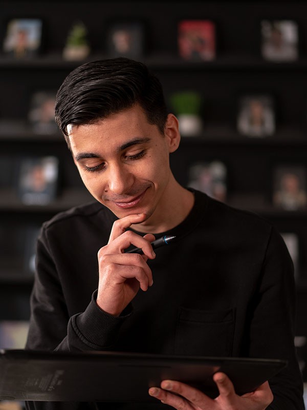 Foto van Taha die denkend naar een tablet kijkt, waarbij op achtergrond vervaagde fotolijstjes te zien zijn